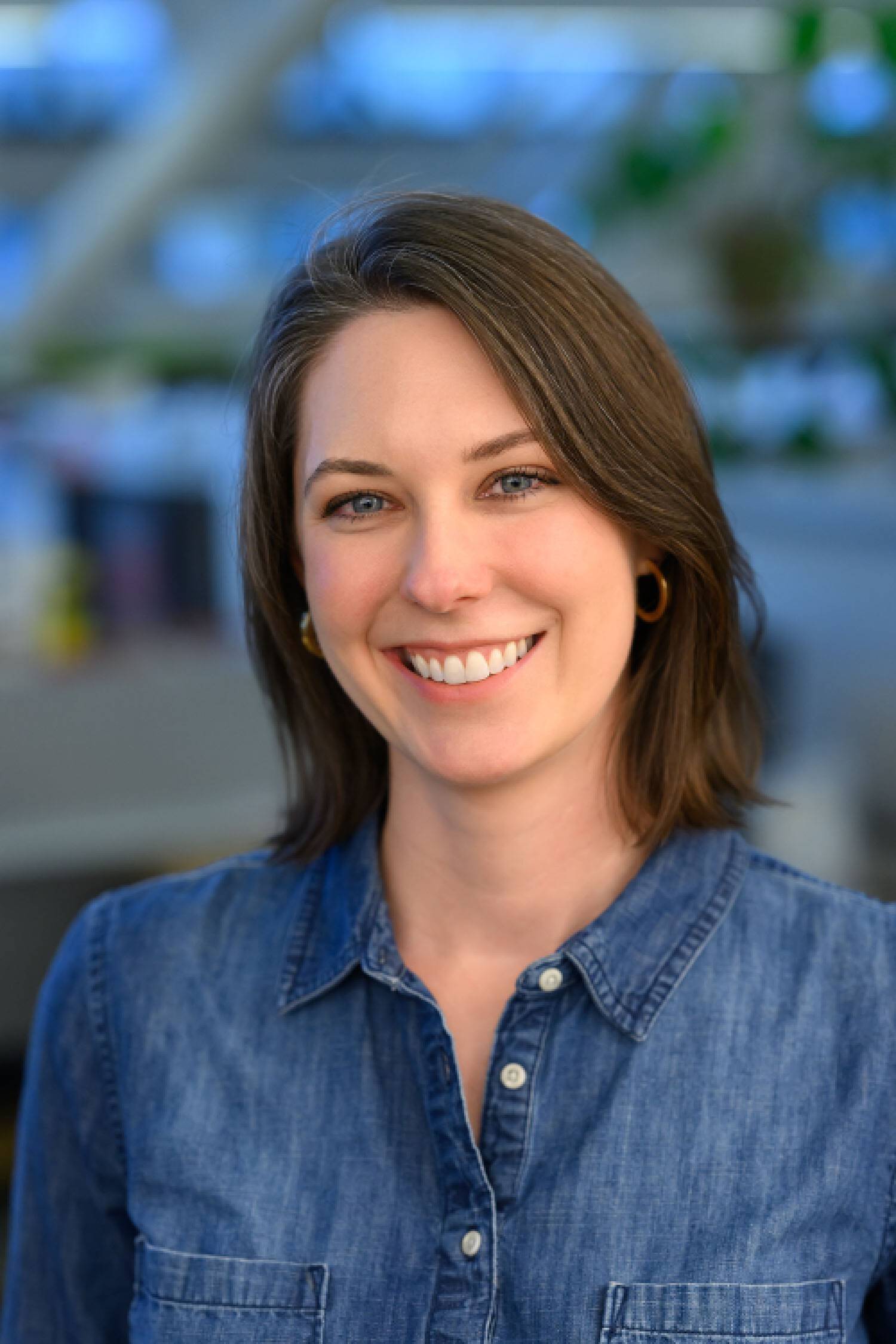 A smiling brown-haired person wearing a denim button down looking at the camera standing in front out a out-of-focus background